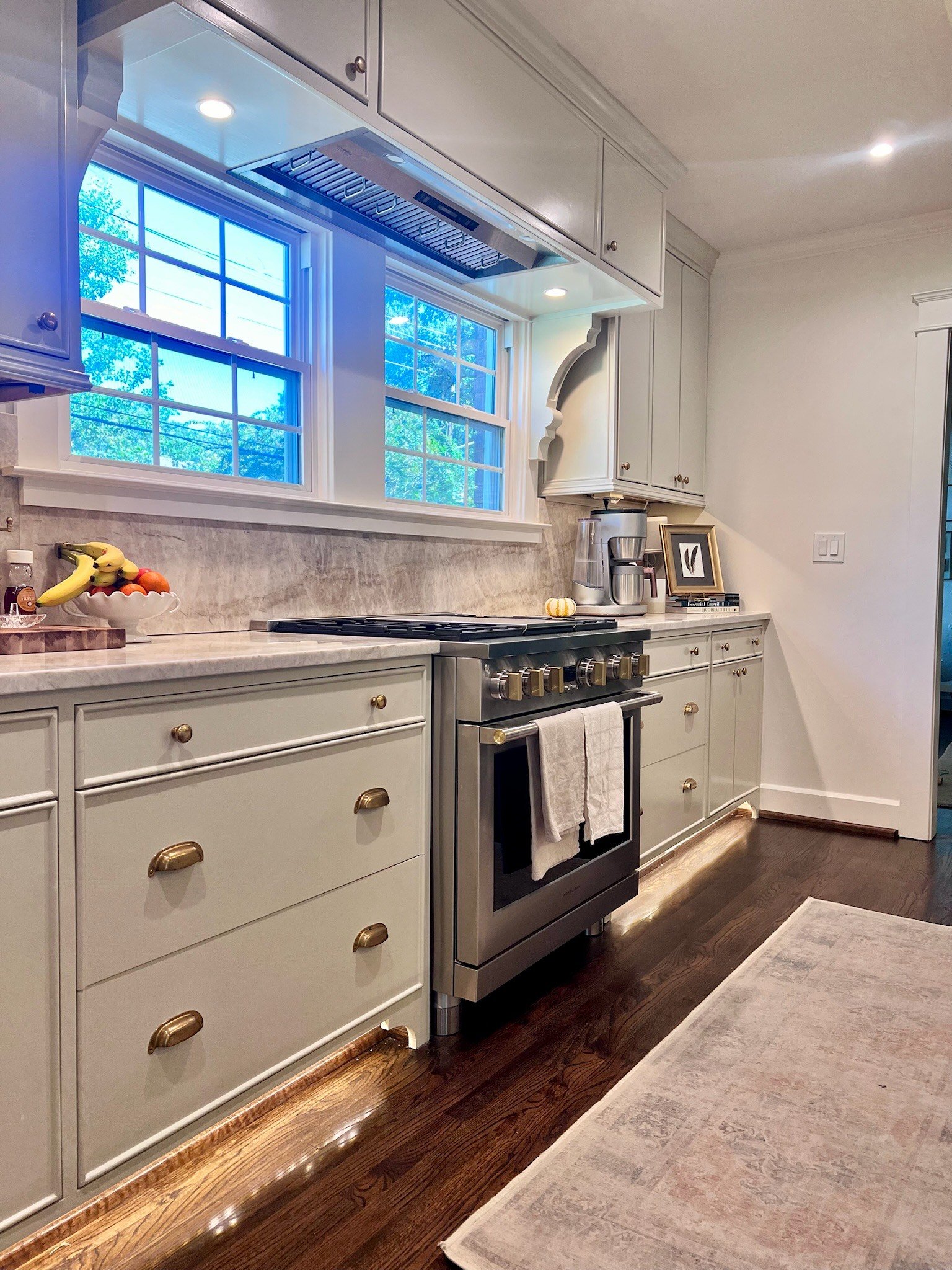 Kitchen range beneath windows with white cabinets, stone counters and brass hardware